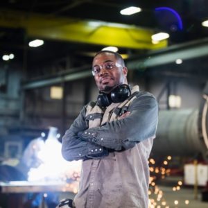 A mid adult African-American man in his 30s working in a metal fabrication factory. He is standing in the foreground with his arms crossed, looking at the camera. Sparks are flying in the background where a worker is welding.
